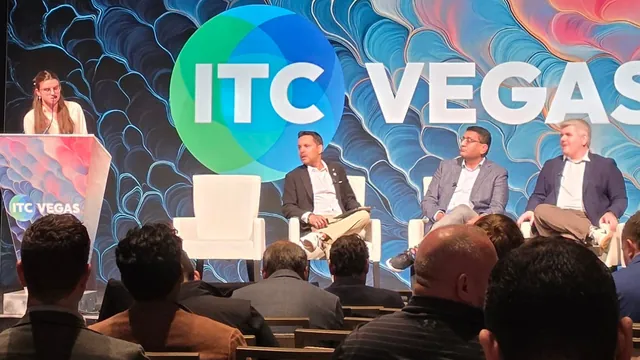 Three panelists seated on white chairs on the ITC Vegas stage while a host speaks at the podium, seen from the audience.